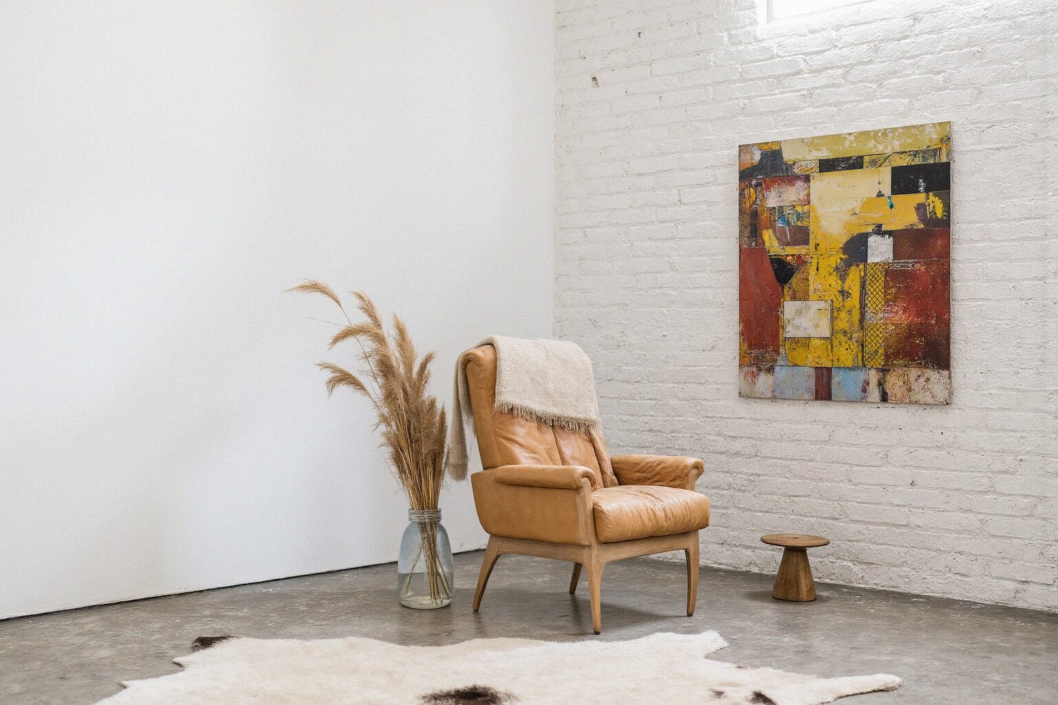 Warm, eclectic boho-Scandinavian corner with white-painted brick, tan leather armchair, cowhide rug and dried grasses